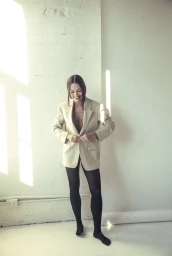 Woman in an oversized beige blazer and black tights buttoning her jacket against a bright studio wall