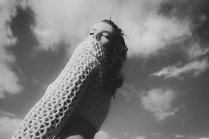 Low angle monochrome photograph of a woman wearing a textured knit sweater against an open sky with scattered clouds.