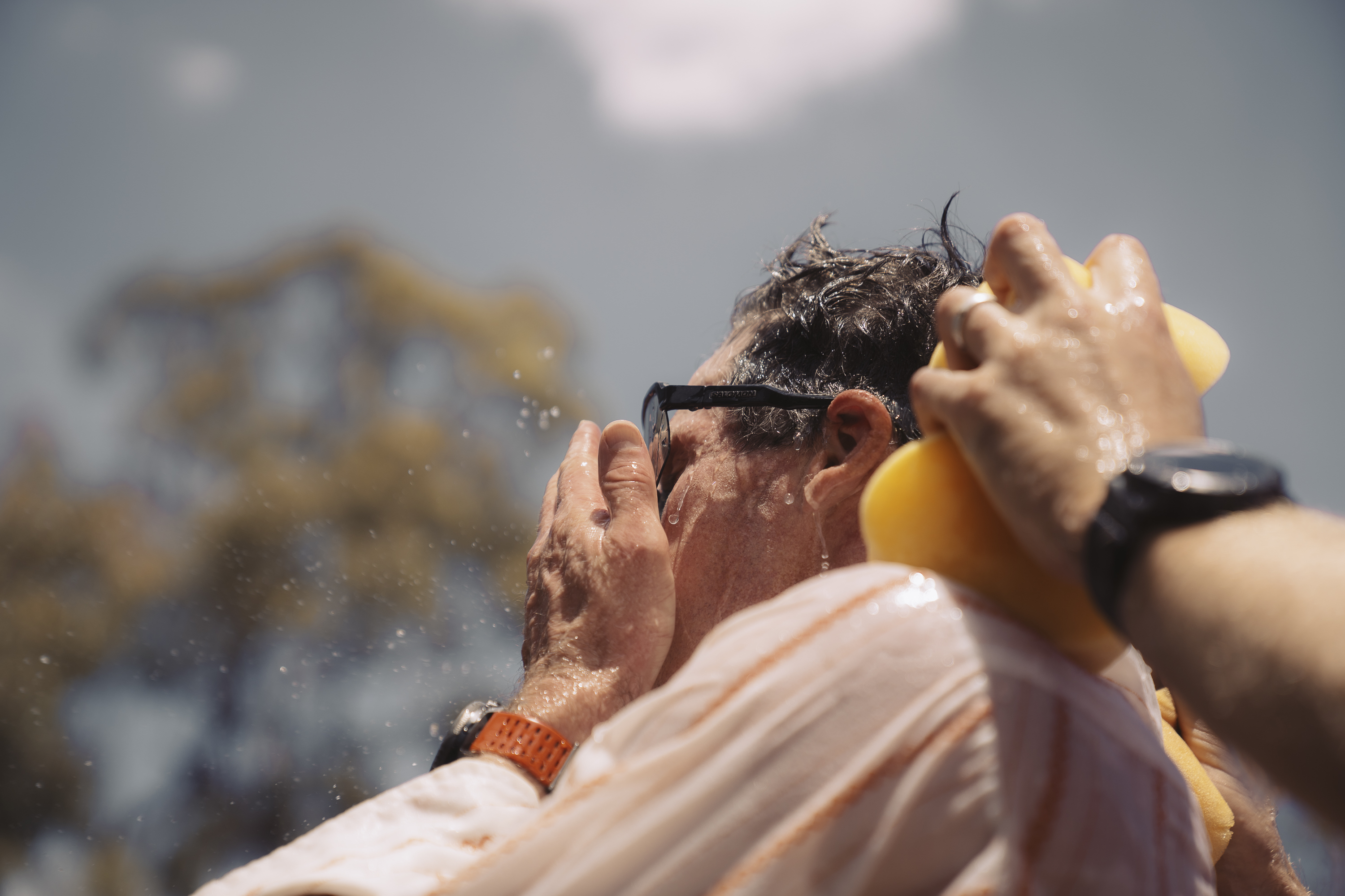 Mature man with sunglasses outdoors being washed down with water and a sponge, wearing an orange wristband and a striped shirt.