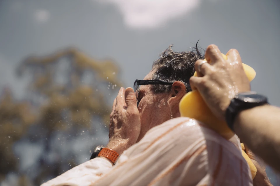 Mature man with sunglasses outdoors being washed down with water and a sponge, wearing an orange wristband and a striped shirt.