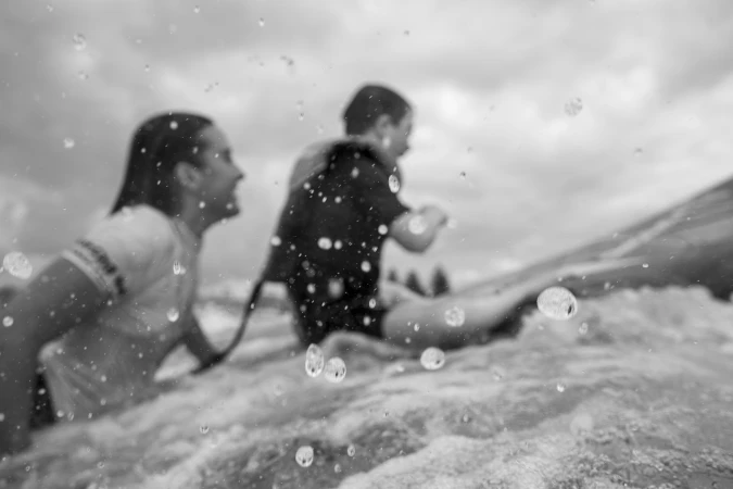 Two people, a woman and a child in a life vest, ride a surfboard on a cloudy day, captured with a close-up and focus on water droplets.