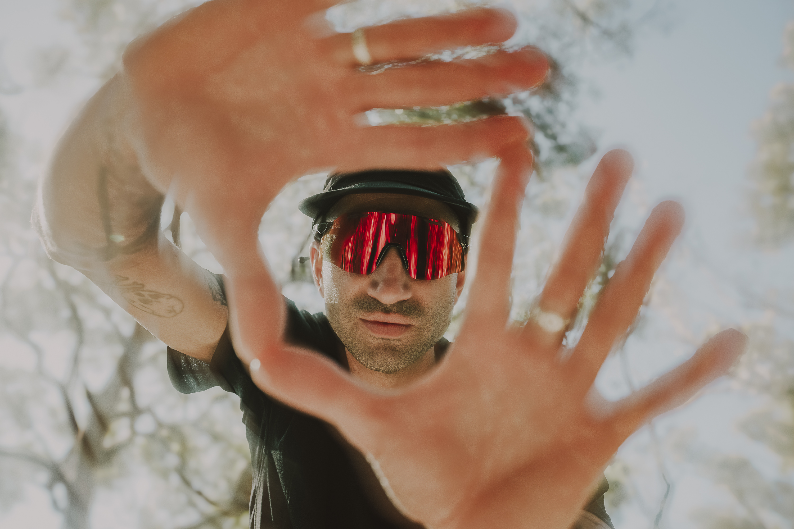 Portrait of a man wearing reflective sunglasses and a hat, holding his hands up to frame his face, against a bright outdoor background.