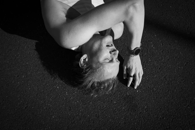 High-angle monochrome portrait of a person lying on dark asphalt wearing a watch.