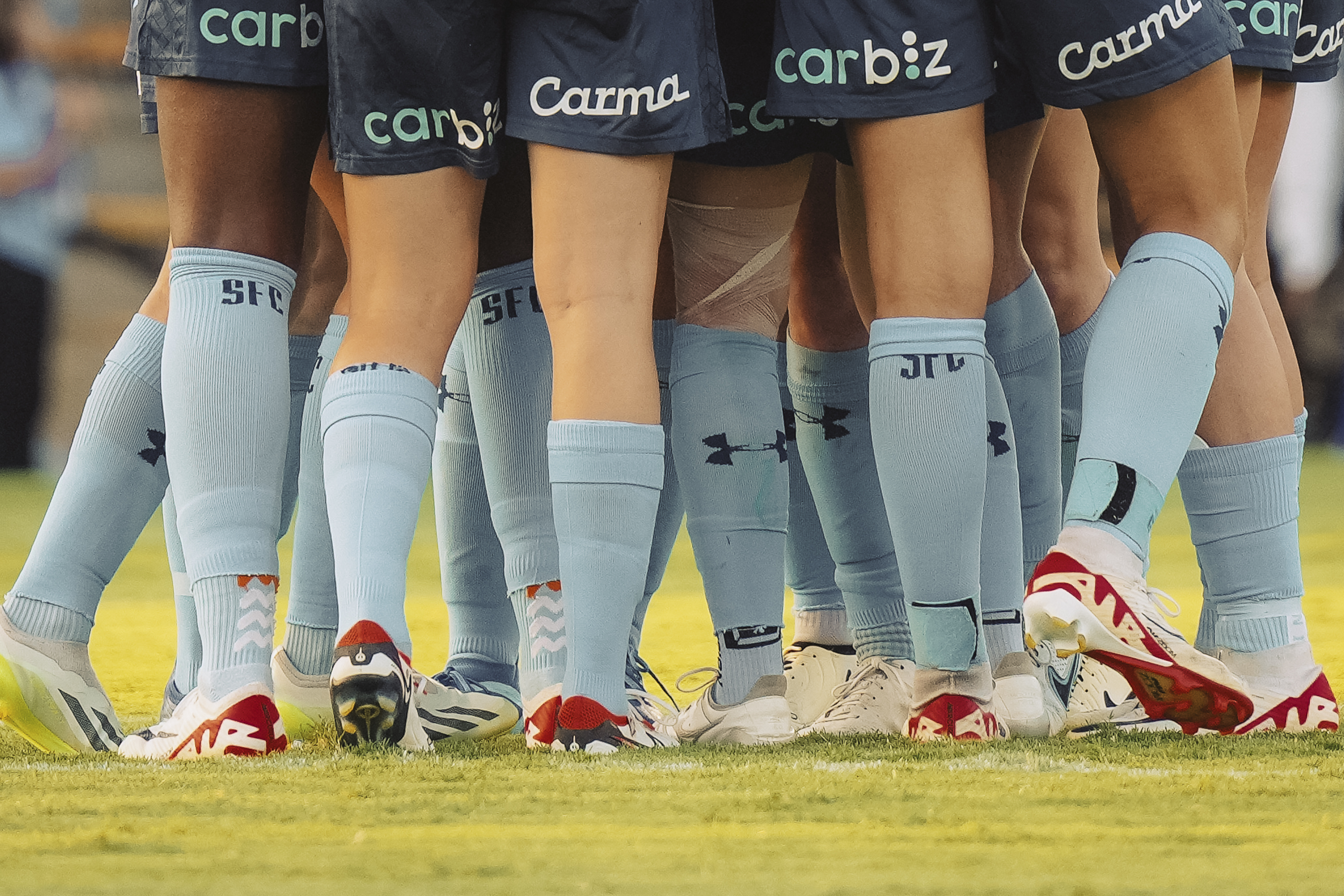 Close-up of a group of soccer players huddled together on a green field, showing their legs, socks, and cleats with branding and logos.