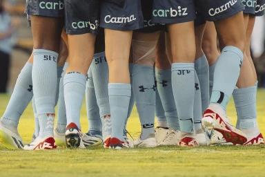 Close-up of female soccer players huddled together in team uniform with blue shorts and light blue socks on a grass pitch.