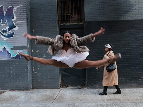 Woman in tutu and fur coat jumps mid-air doing splits in an urban street setting with graffiti wall and a pedestrian in the background.