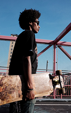 Young adult man with dark curly hair wears sunglasses, carries skateboard on urban bridge with blue sky, stands in profile view.