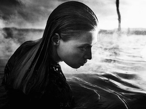 Monochrome portrait of a woman in a body of water, with wet hair and a contemplative expression, captured in a close-up studio shot.