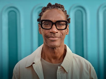 Portrait of a Black man with short locs and black thick-rimmed glasses wearing an off-white jacket, against a turquoise arched wall.