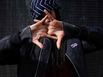 Portrait of a person wearing a dark quilted bomber jacket and striped headwrap making a hand sign
