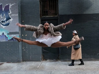 Black ballerina in a white tutu and fur coat leaping mid-air in front of a dark brick wall on a city street