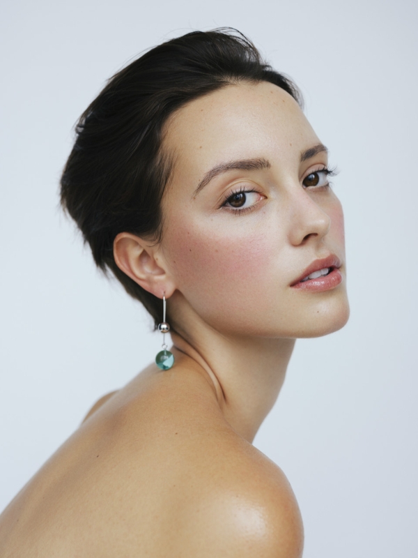 Close-up portrait of a young woman with short brown hair wearing a dangling teal earring against a plain white background.