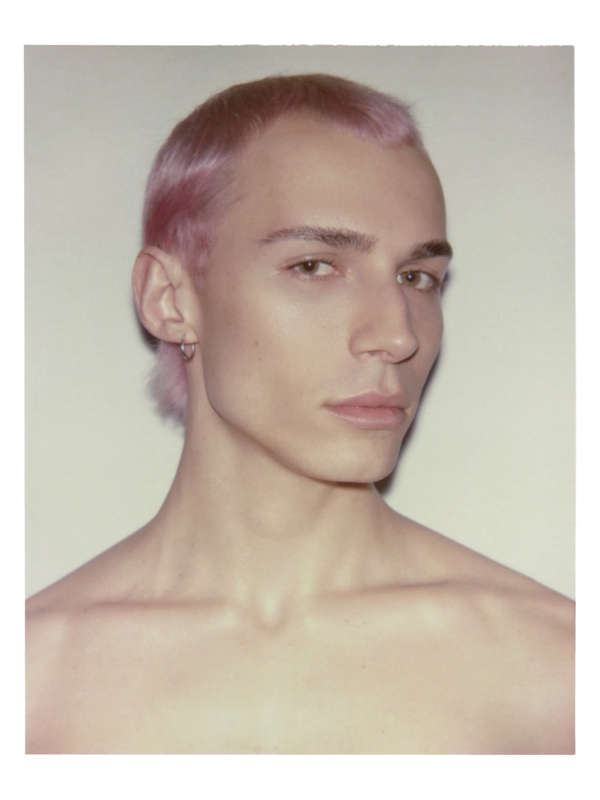 Close-up portrait of an adult male with pink hair and bare shoulders against a neutral background, showcasing his features and unique hair color.