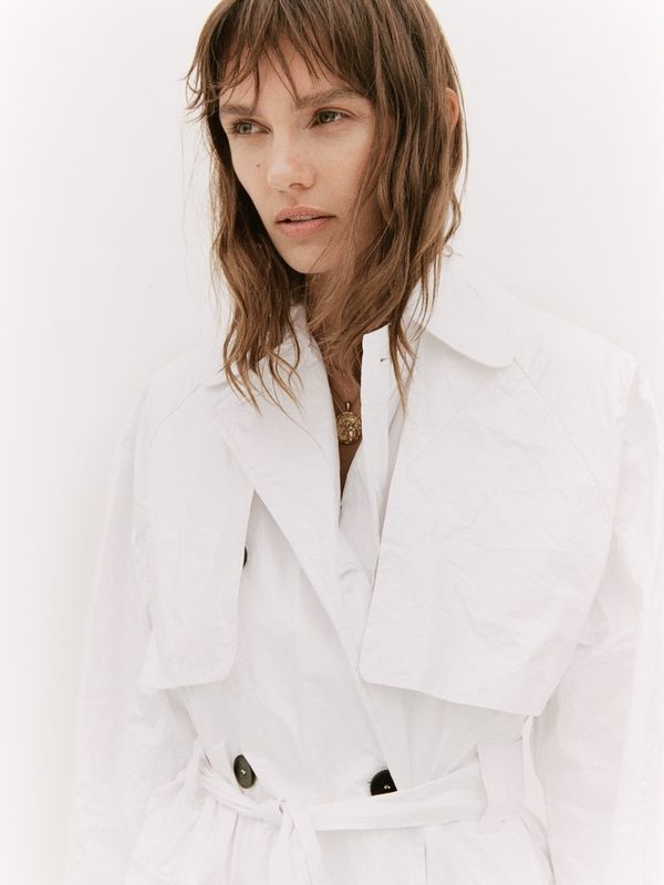 A fair-skinned adult female model wearing a white trench coat poses against a white background in a fashion editorial, with medium shot.