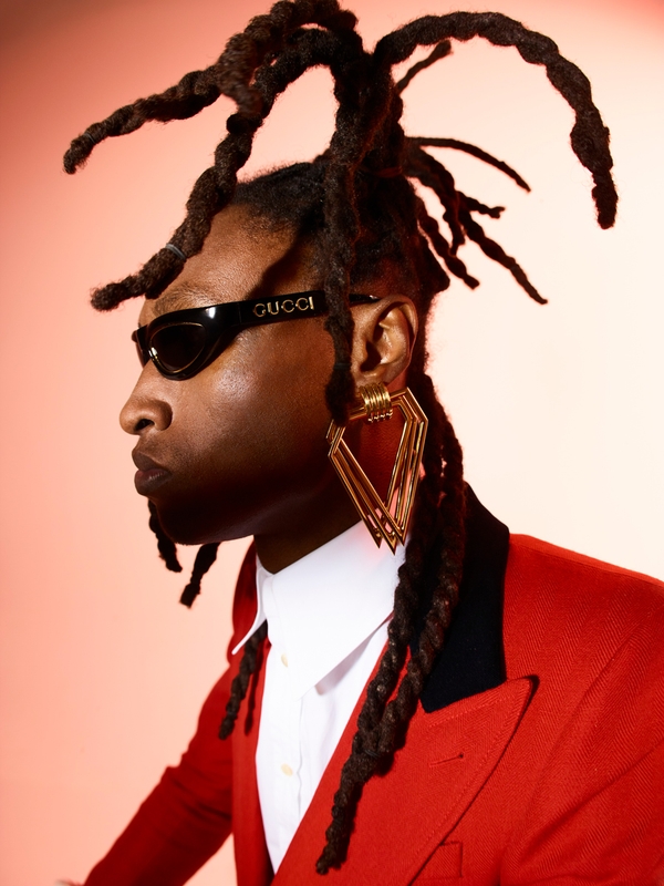 Headshot of Black male model in a red suit and black sunglasses with Gucci logo, and long dreadlocks, against a gradient peach background.