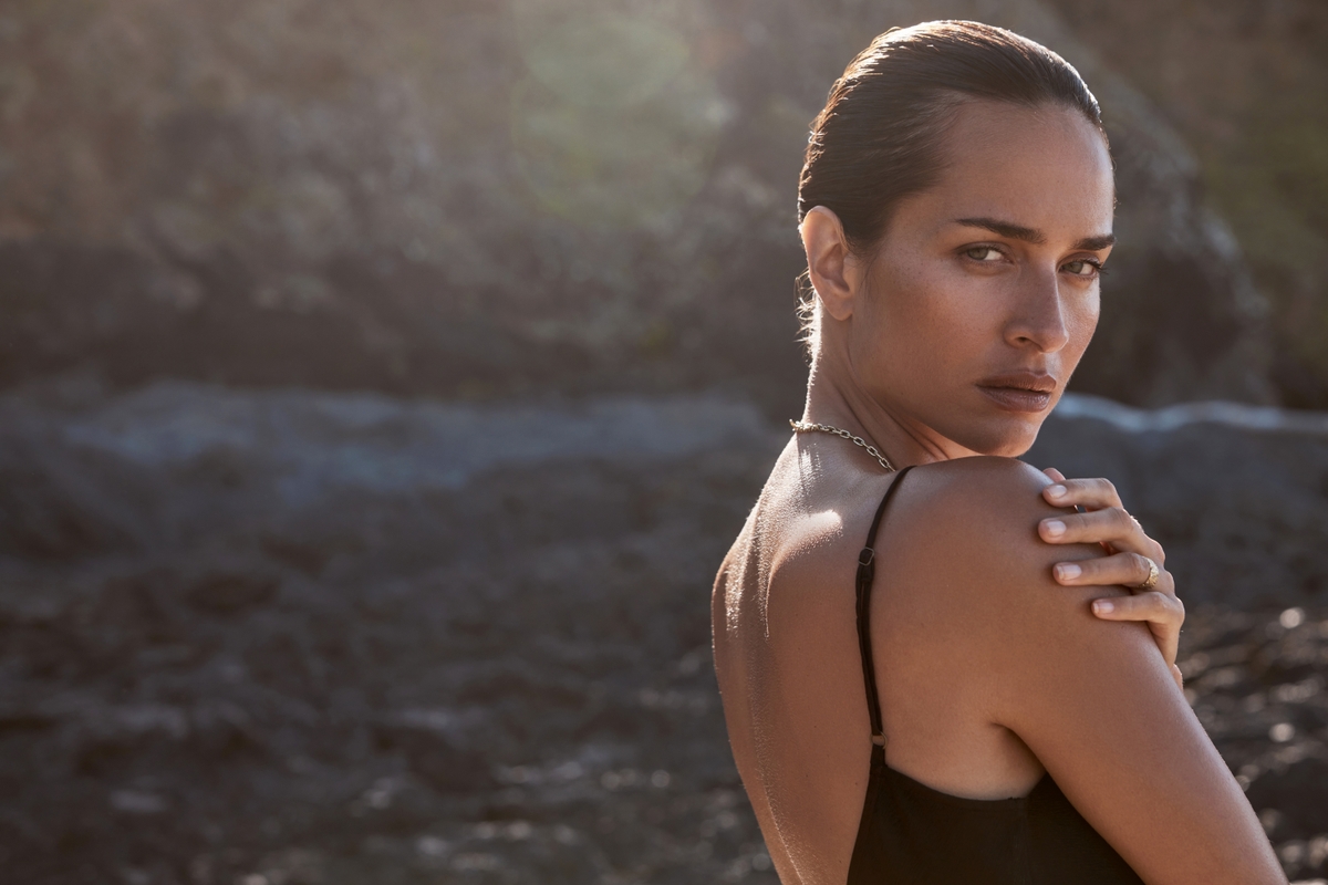 Adult woman gazes over her shoulder, wearing a black top and necklace, set against a blurry, neutral rocky outdoor backdrop.