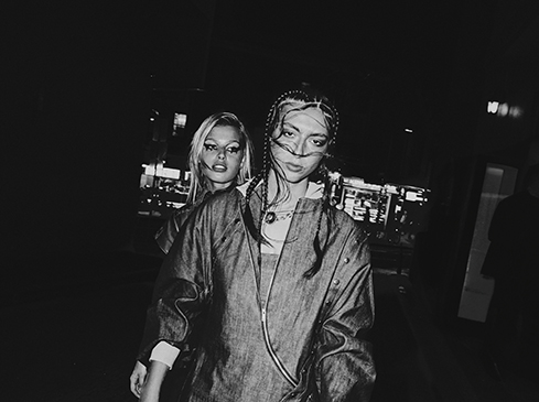 Two women pose in denim jackets against an urban backdrop at night, illuminated by city lights, rendered in monochrome for a stylish, gritty aesthetic.