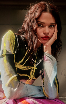 Portrait of a woman with curly hair wearing a colorful long-sleeve top and red lipstick, posing with her hand on her face.