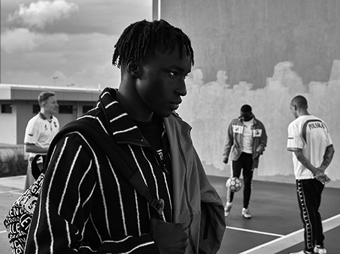 Black and white shot features a young Black man with dreadlocks in a striped jacket, with other men playing soccer in the background.