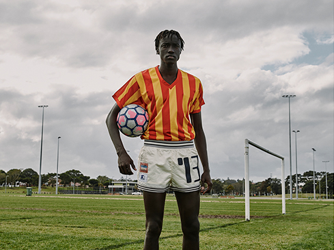 Athletic man in sports jersey and shorts poses with soccer ball on field under cloudy sky; goal and field lights in background.