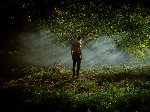 Bare-chested adult male stands in a forest with light rays and green foliage, creating a dramatic, natural scene.