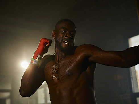 An adult male boxer with dark skin, wearing red hand wraps and a necklace, stands in a boxing pose with a determined facial expression.