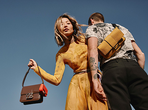 Two fashion models pose with handbags against a clear blue sky. One wears a tan dress, the other a graphic tee and black pants.