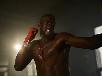 Sweaty black male boxer with red hand wraps throwing a punch in a dim indoor training space photographed by Lester Jones