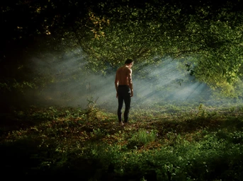 Shirtless man standing in a sunlit forest clearing with mist filtering through the dense green trees captured by Lester Jones.