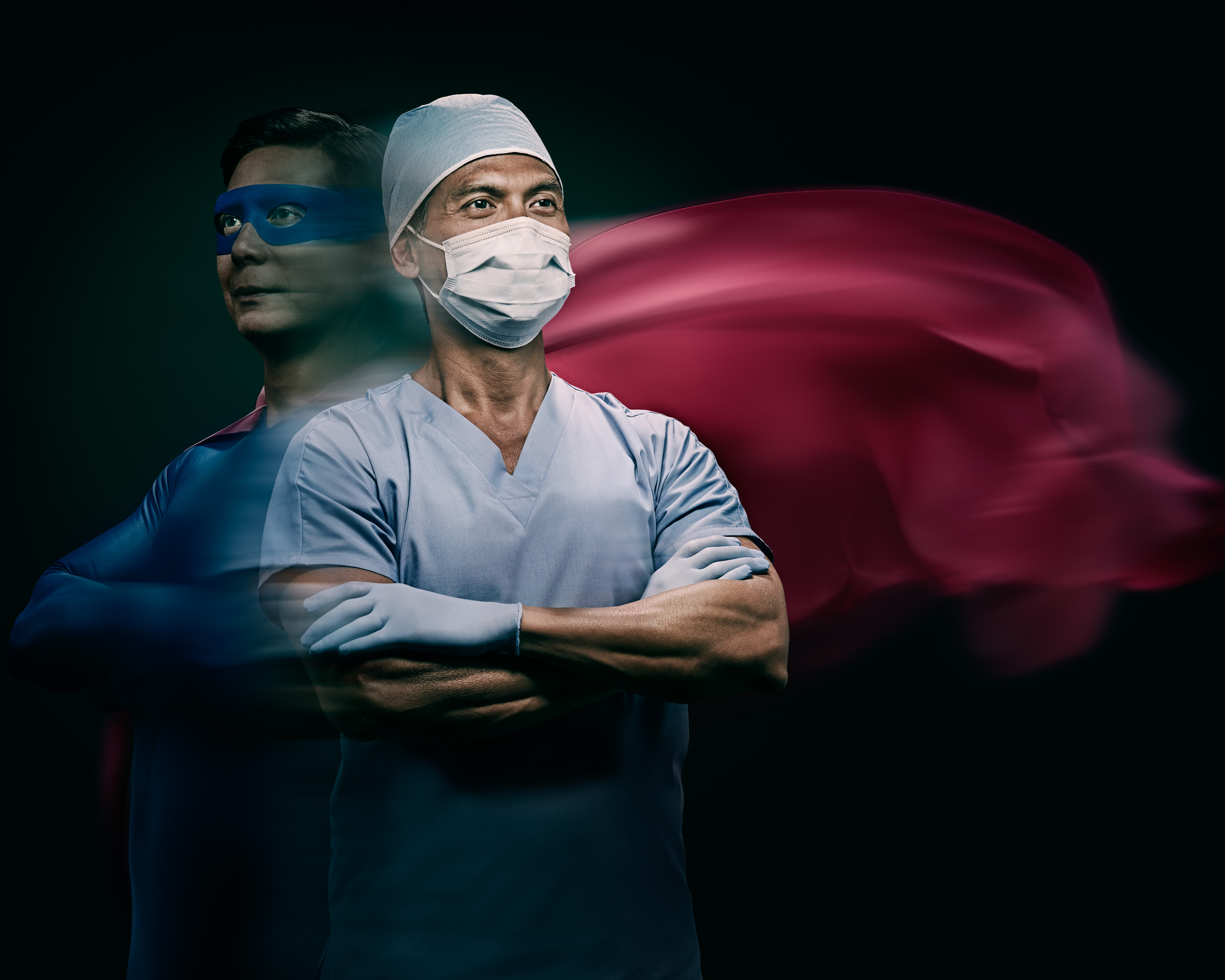Heroic healthcare worker with cape and medical mask stands confidently with arms crossed in a studio setting, showcasing strength and dedication.