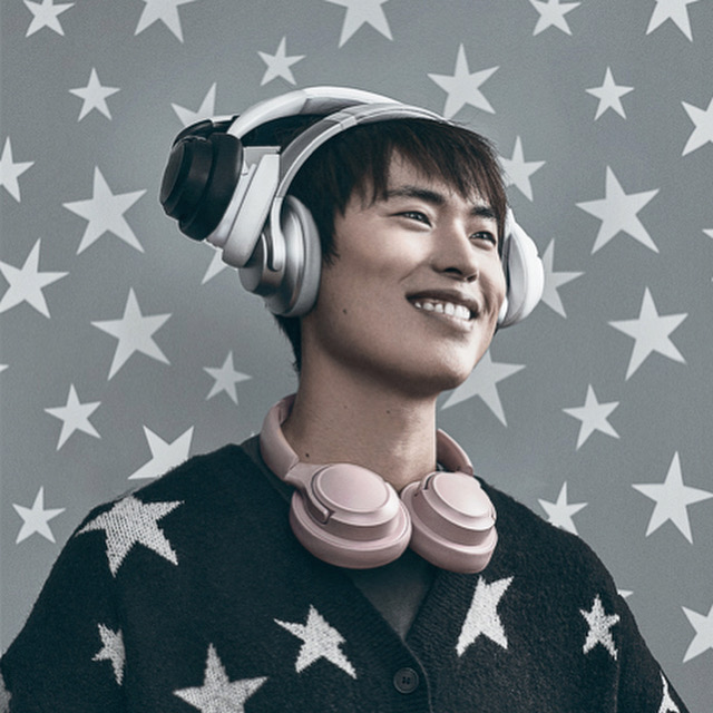 Young man smiles wearing white and pink headphones, adorned in a star-patterned cardigan against a backdrop of star shapes.