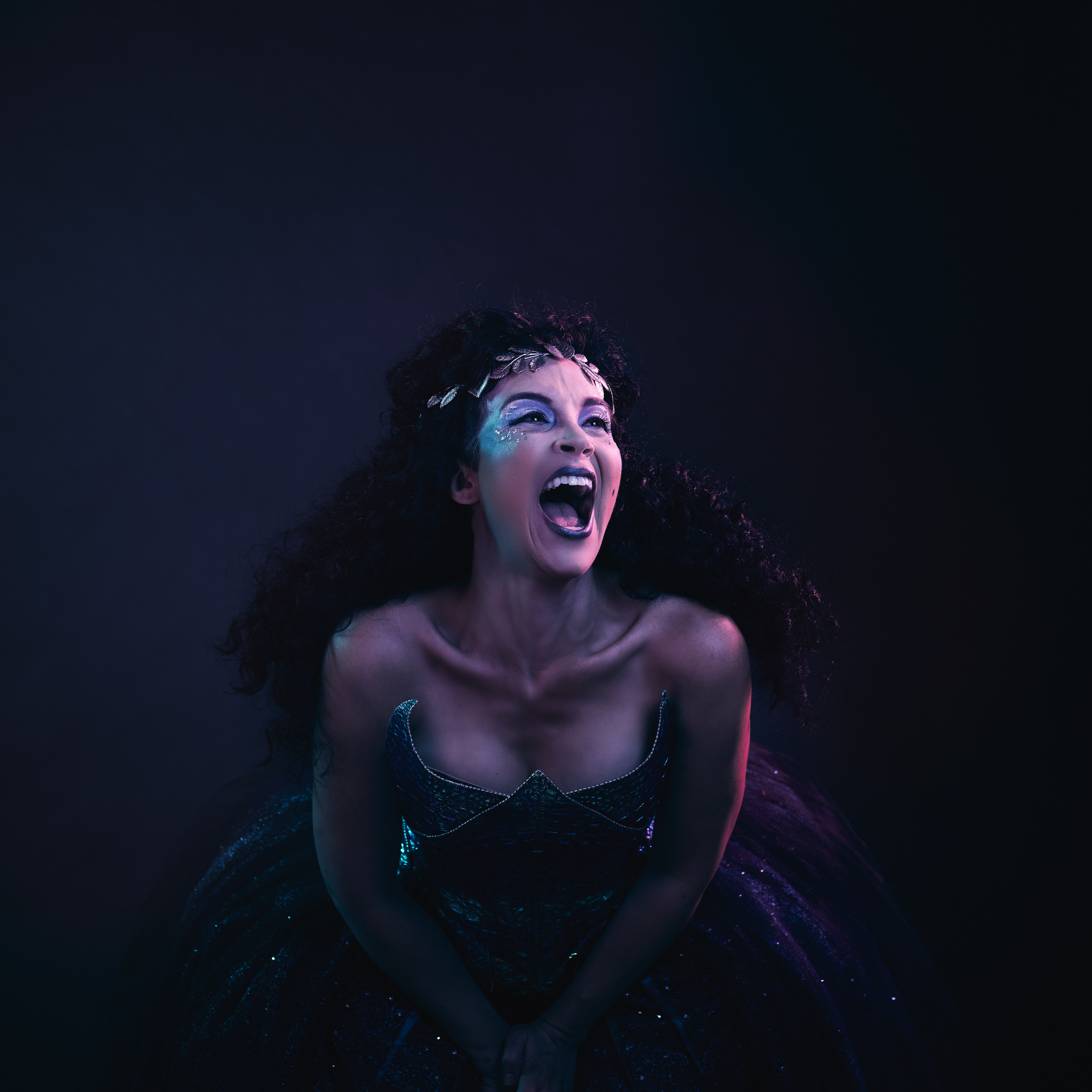 Portrait of a woman in a blue gown with glitter makeup shouting with her mouth open in front of a dark background in studio lighting.