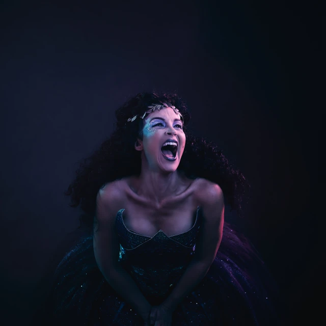 Portrait of a woman in a blue gown with glitter makeup shouting with her mouth open in front of a dark background in studio lighting.