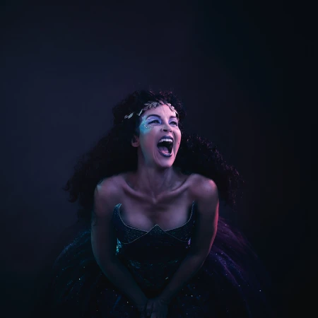 Portrait of a woman in a blue gown with glitter makeup shouting with her mouth open in front of a dark background in studio lighting.