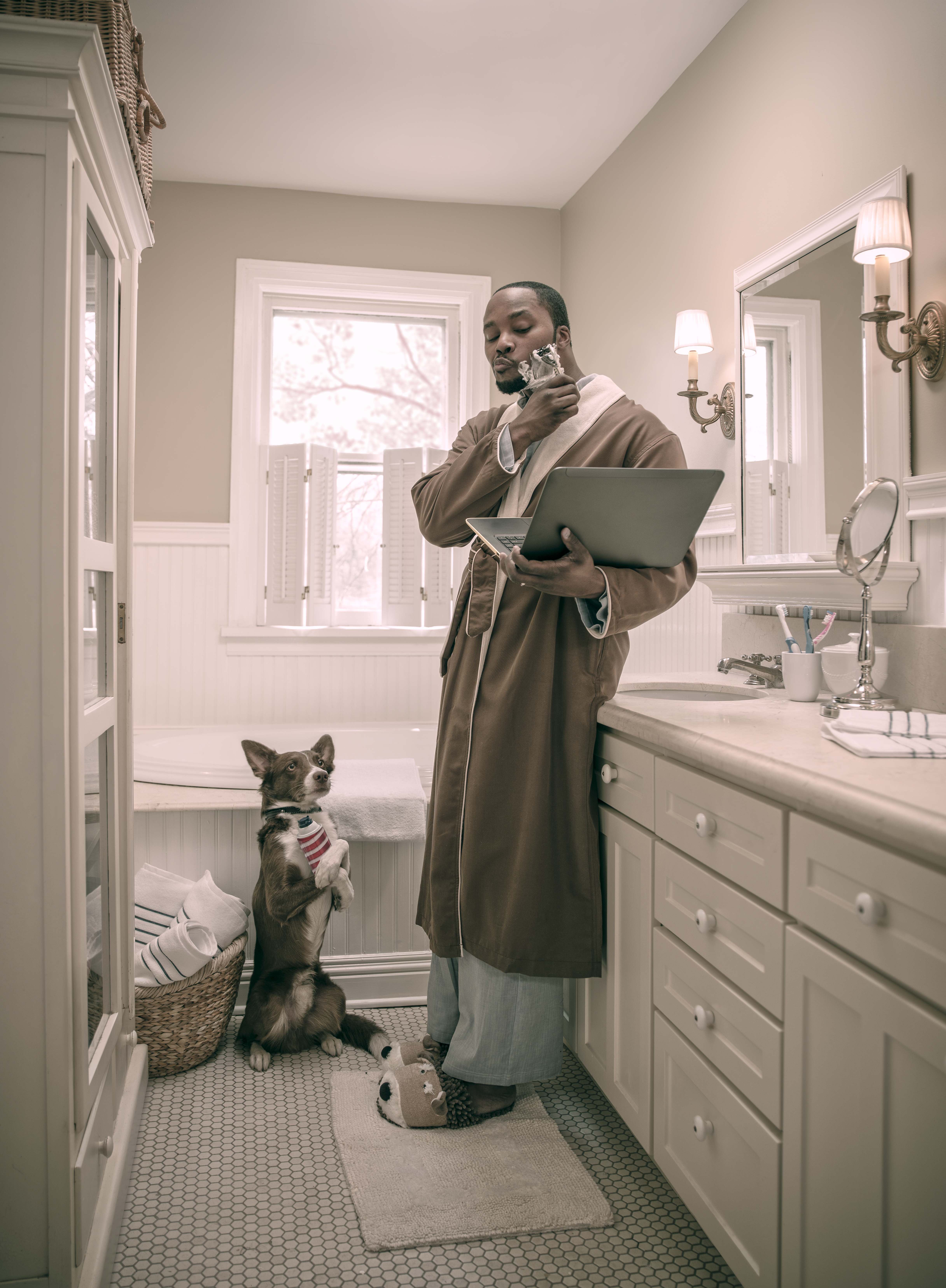 An African-American man in a robe shaves while using a laptop in a bright bathroom, with a dog standing on its hind legs nearby.