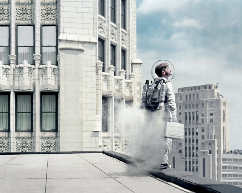Man in space suit with jetpack on city rooftop, holding briefcase, looking up at the sky with building in the background.