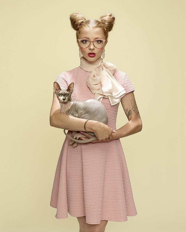 Woman with space buns and glasses holds a hairless cat in a studio portrait against a light-yellow background, wearing a pink dress.