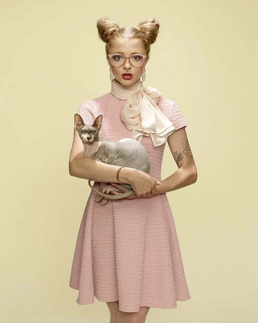 Woman with space buns and glasses holds a hairless cat in a studio portrait against a light-yellow background, wearing a pink dress.