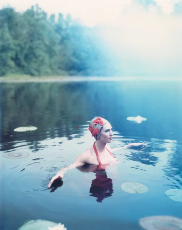 Woman in a red swimsuit and floral bathing cap in a tranquil blue lake surrounded by lily pads and green trees with a misty backdrop.