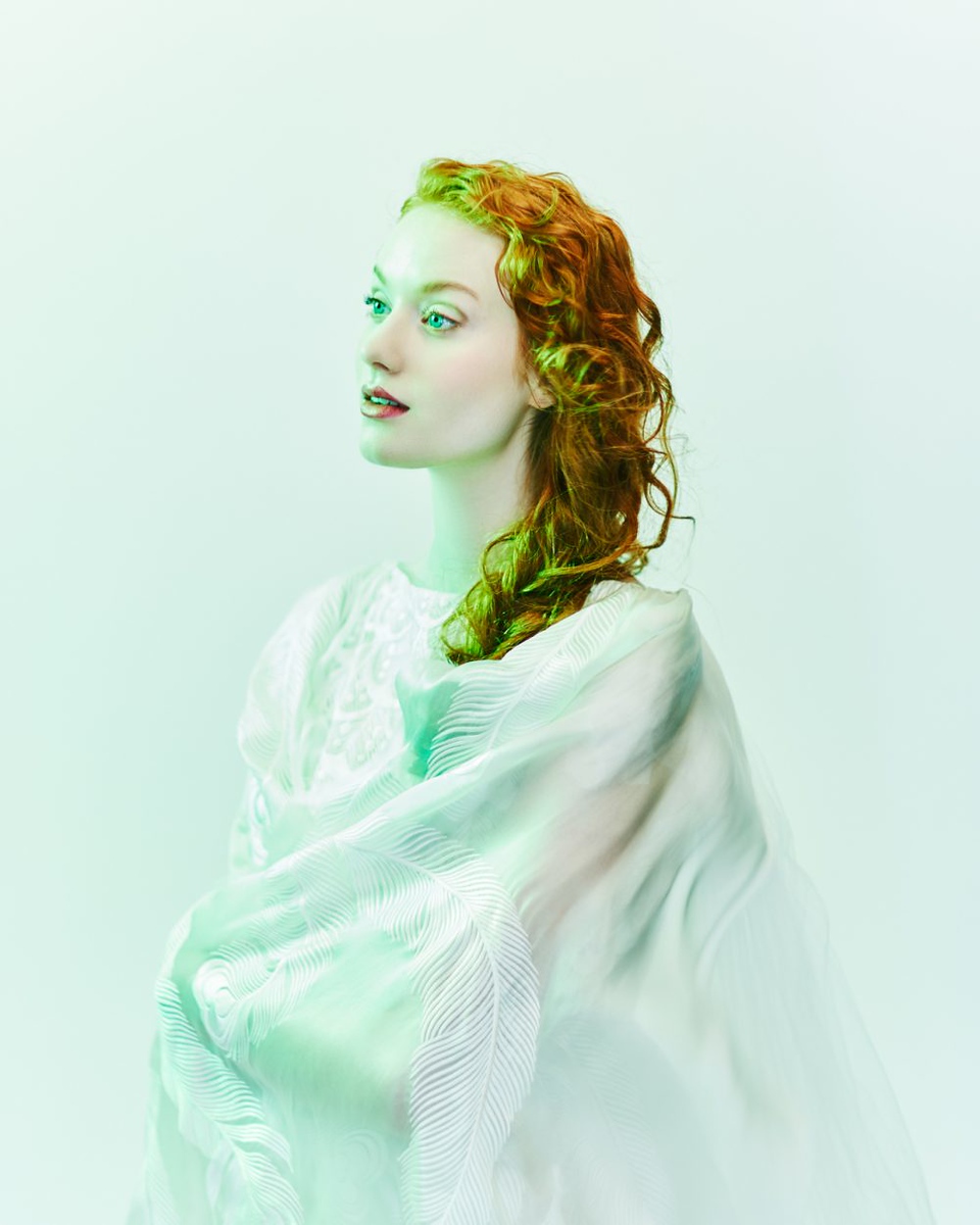 Woman with red curly hair gazes off-camera, draped in sheer white fabric, illuminated by soft, diffuse lighting in a portrait.