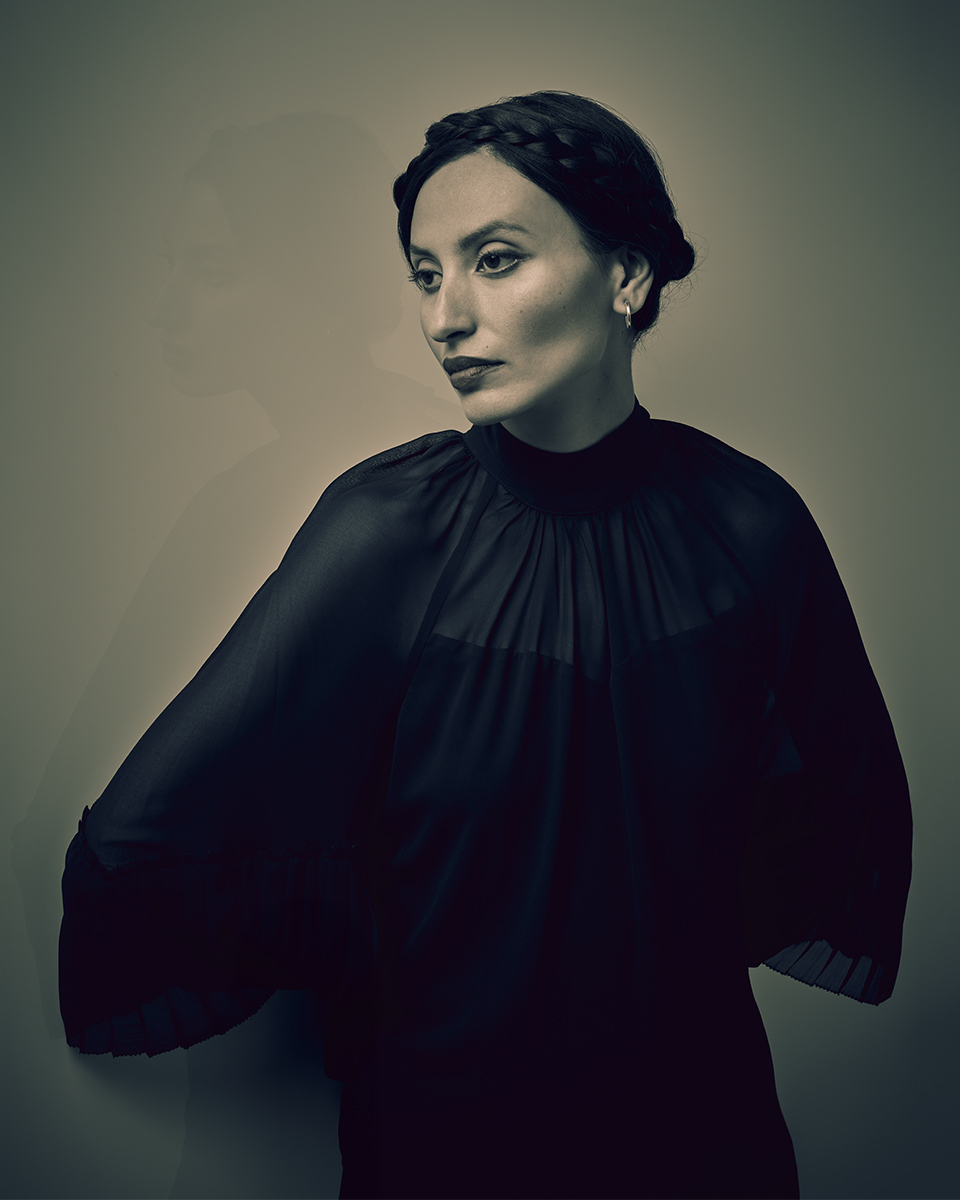 Portrait of an adult female with braided hair in a dark, elegant dress against a plain background, looking off-camera.