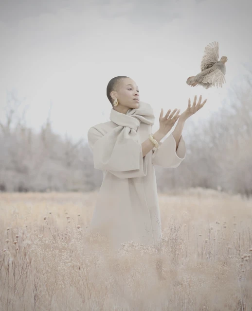 Woman in a cream-colored coat releases a bird into the air in a hazy field. The bird flies above outstretched hands.