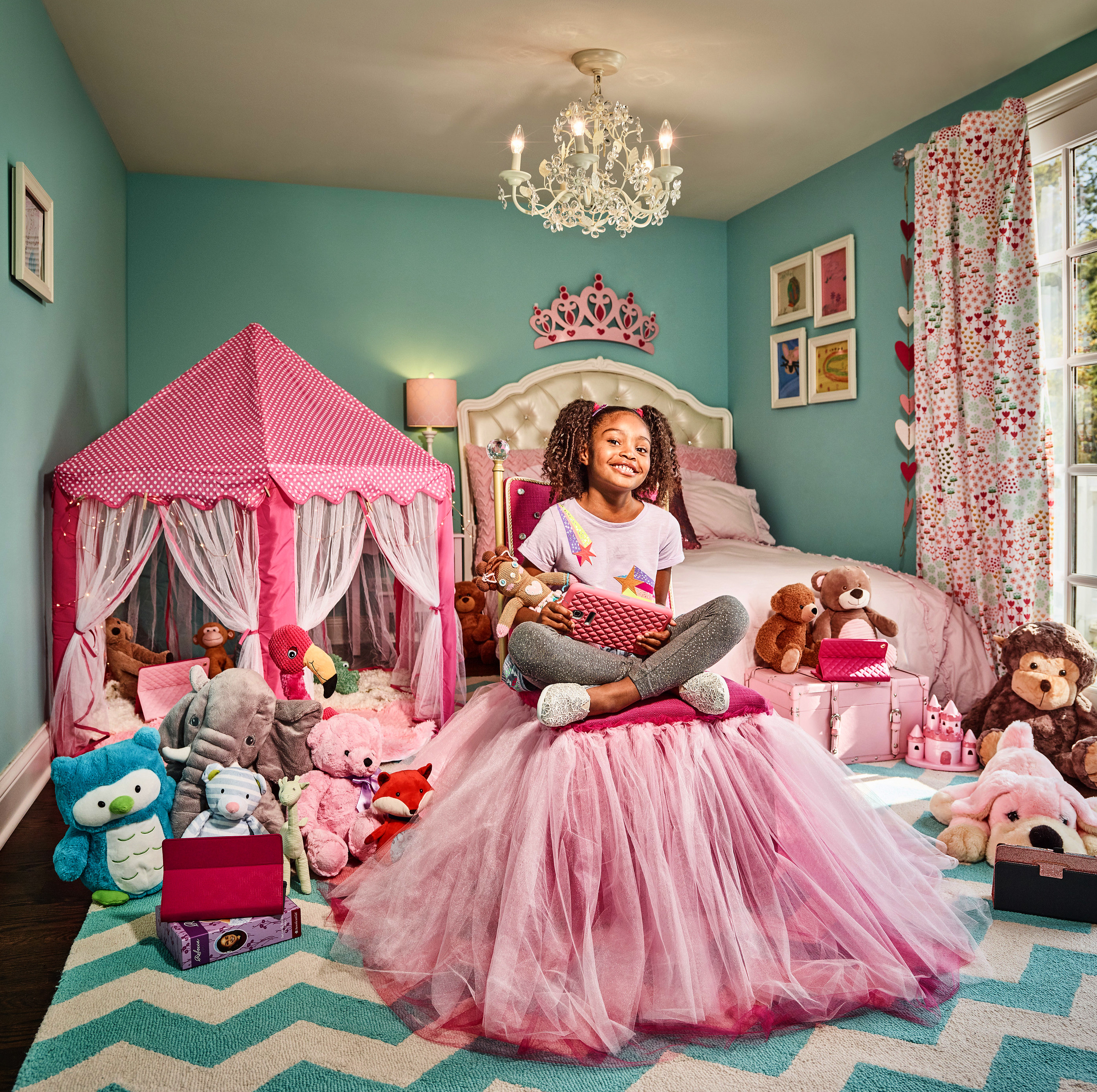 Young girl sitting cross-legged on a pink tulle dress in a turquoise bedroom with a toy tent and teddy bears, smiling at the camera.