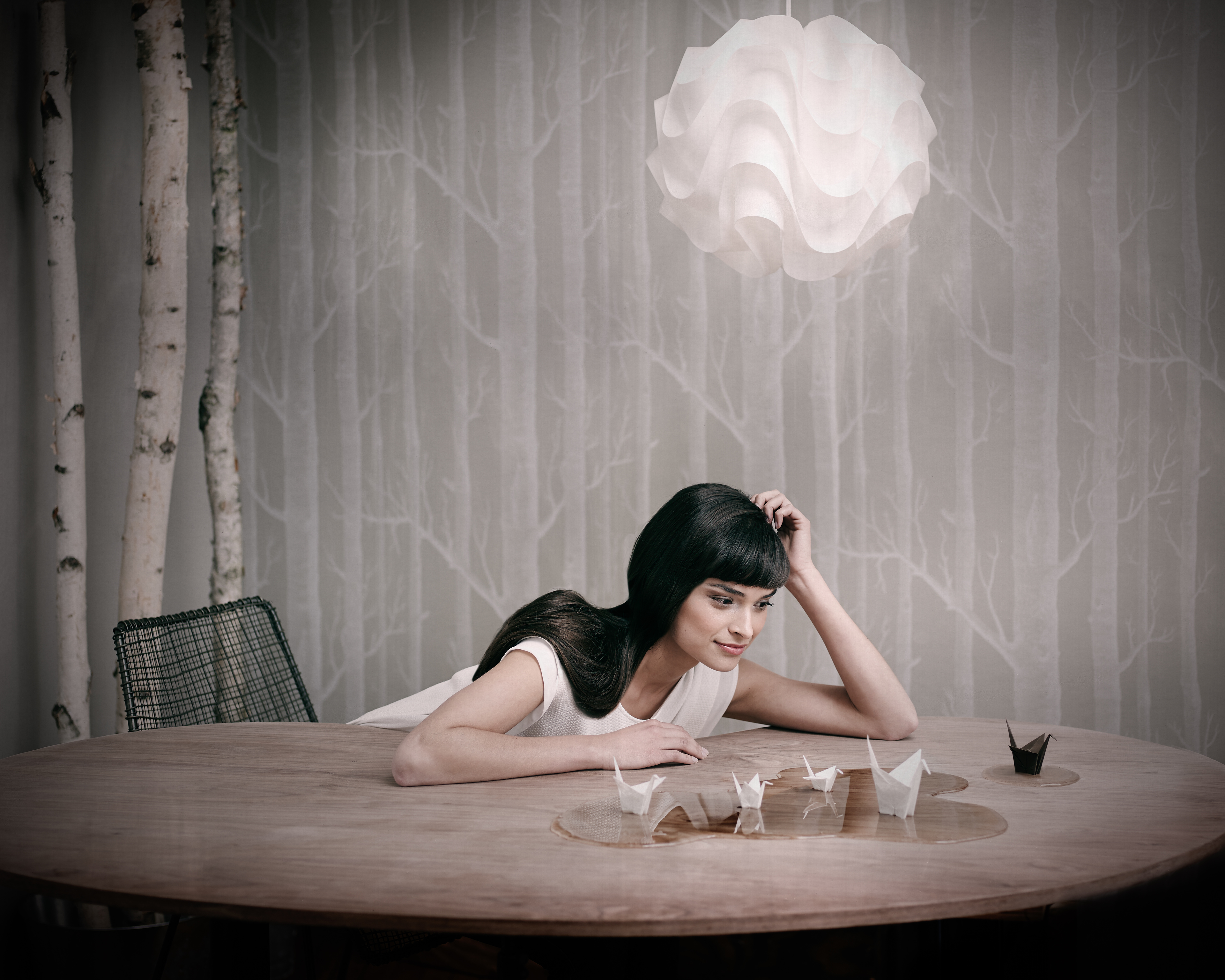 Young woman sits at a table with origami cranes reflected in spilled water, with a paper lamp and birch-tree-patterned wallpaper backdrop.