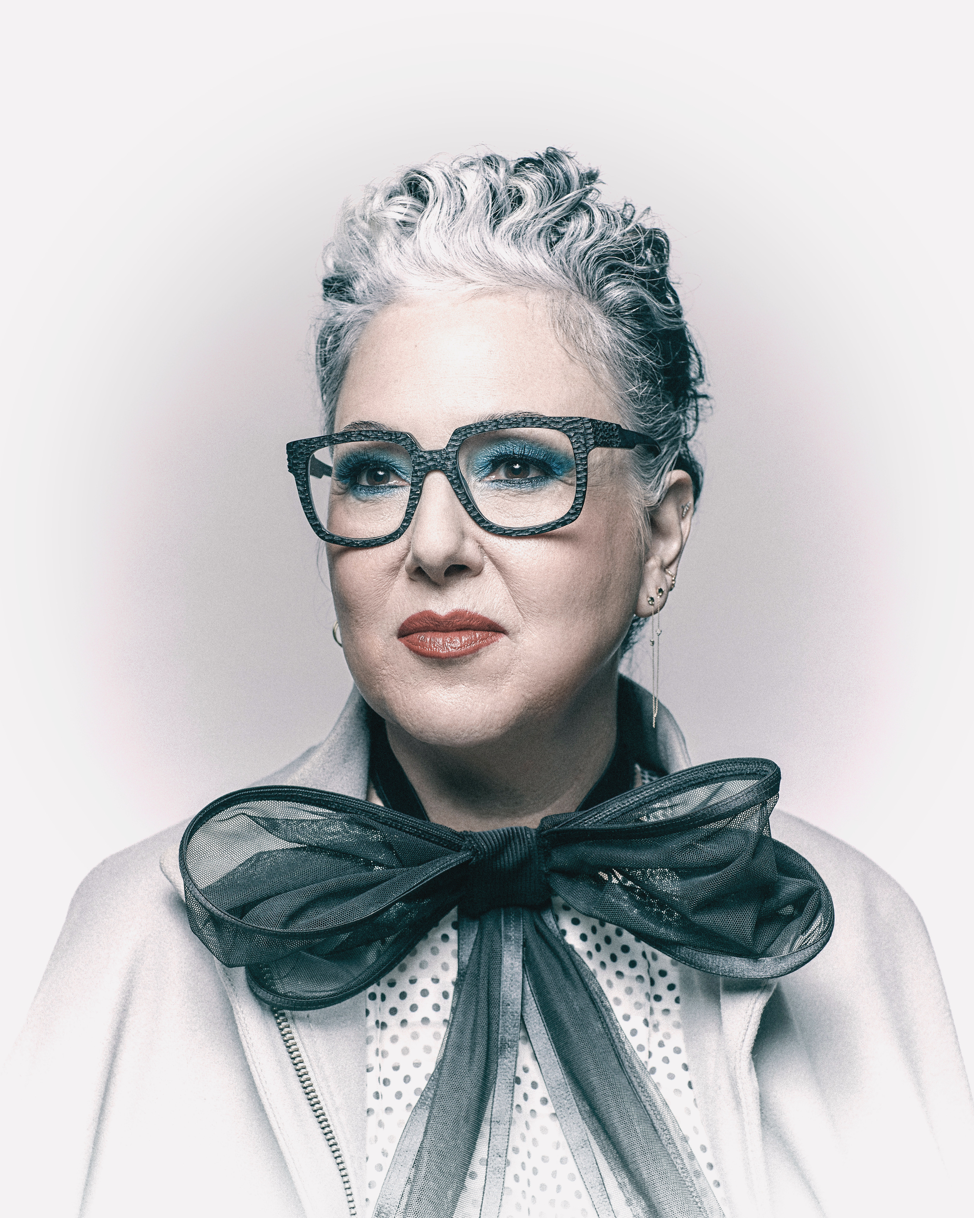 Portrait of an adult woman with grey hair wearing eyeglasses and a white coat against a light background in a photo studio.