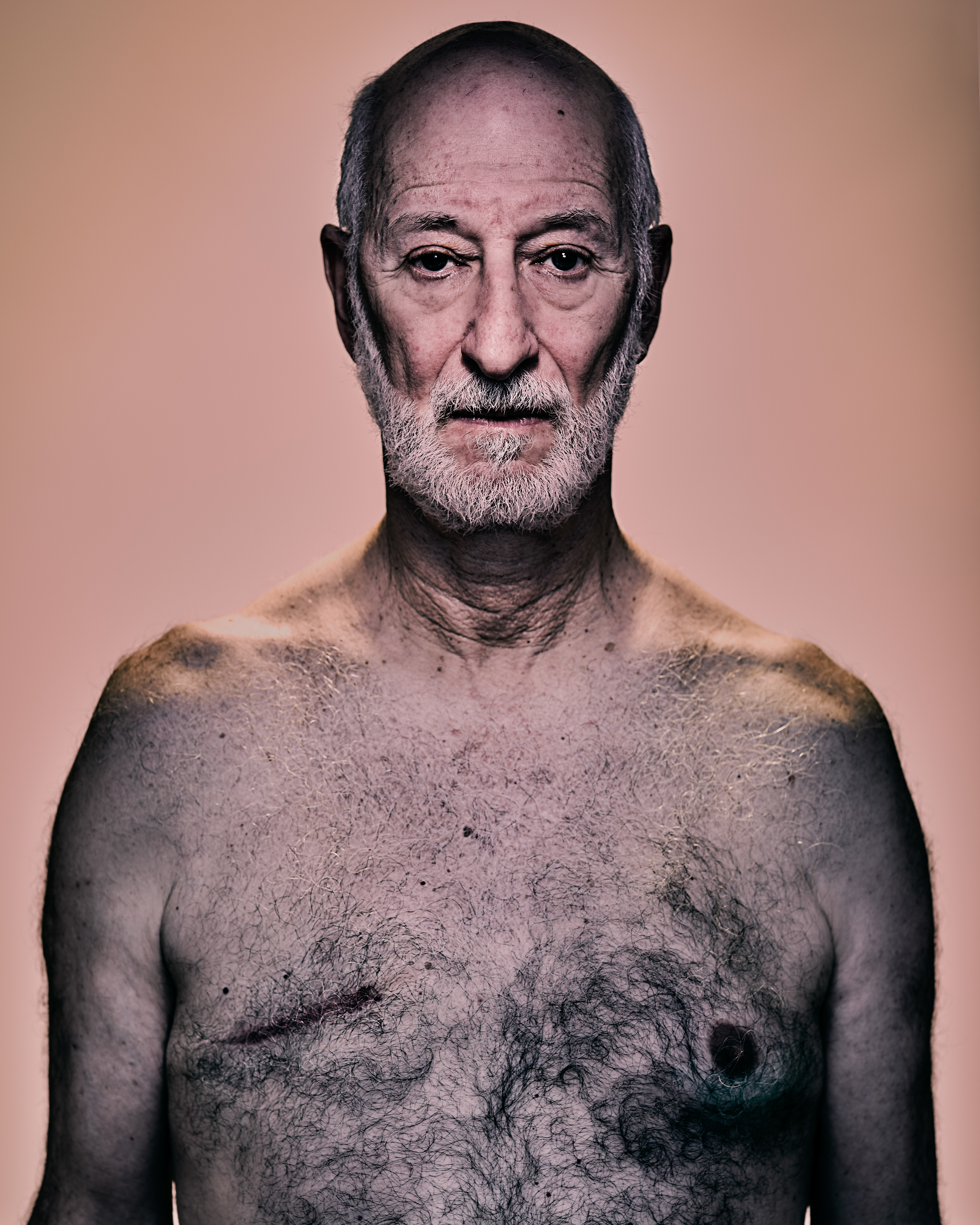 Close-up portrait of a mature adult man with white beard and bare chest against an orange background, showcasing his weathered skin.
