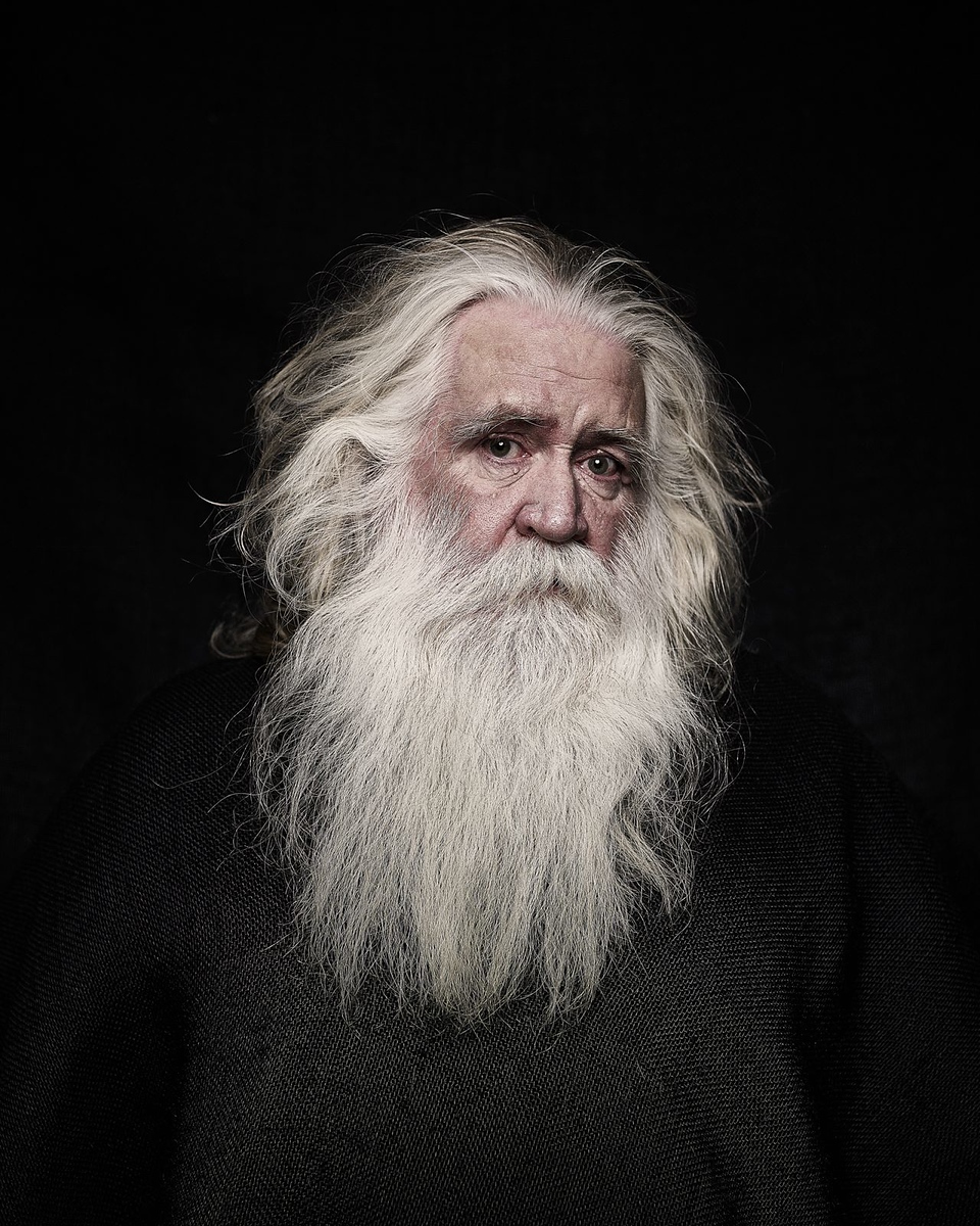 Portrait of an elderly man with long, flowing white hair and beard, wearing a dark sweater against a stark black background.