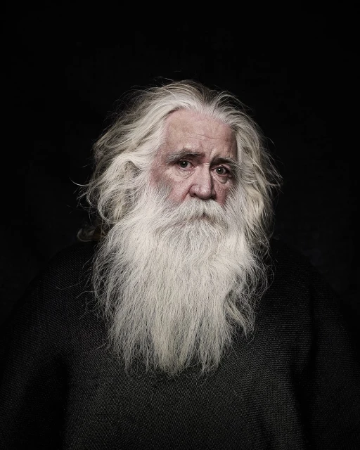 Portrait of an elderly man with long, flowing white hair and beard, wearing a dark sweater against a stark black background.