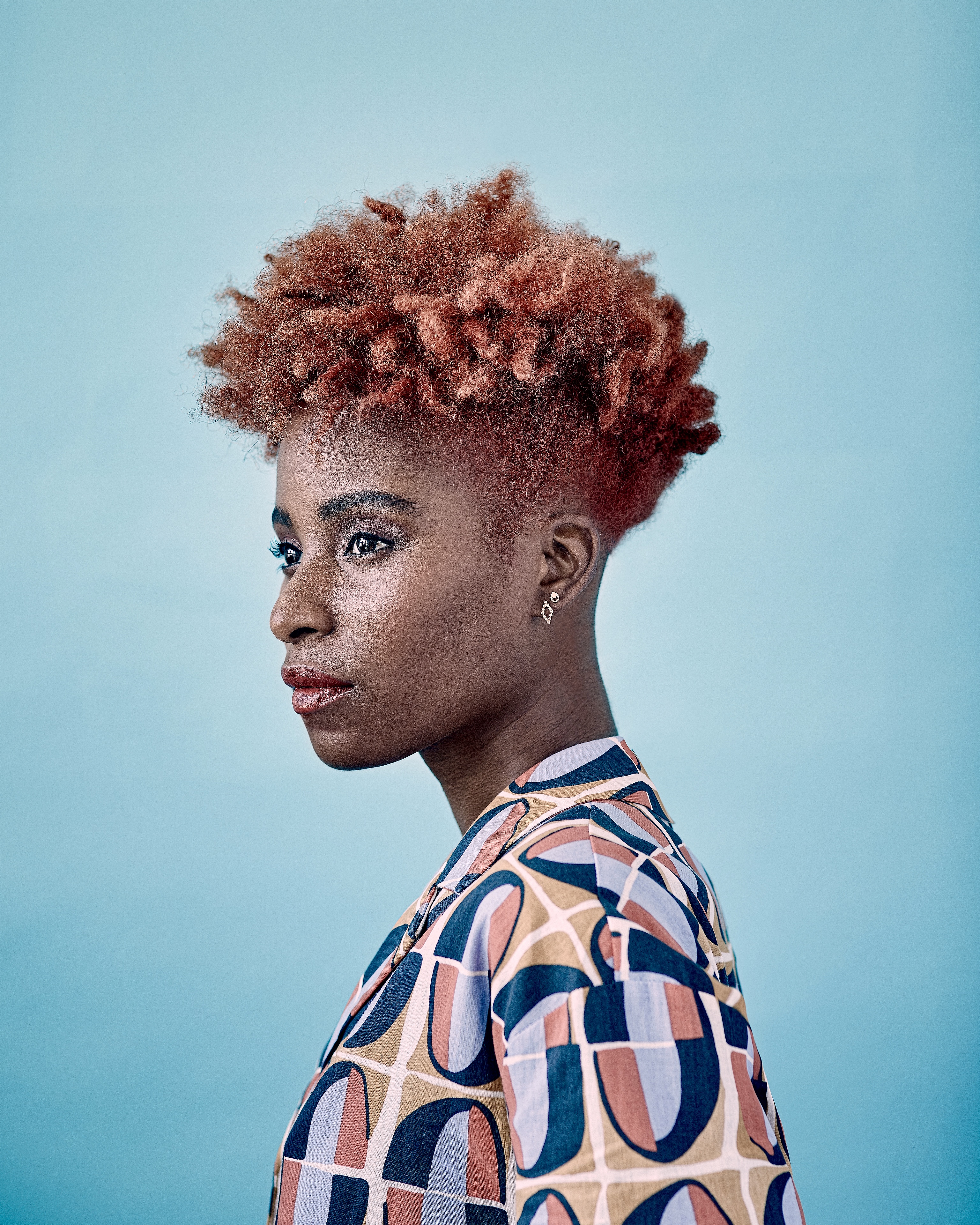Stylish portrait of a Black woman with short, copper-colored curly hair, wearing a geometric-patterned jacket against a light blue background.