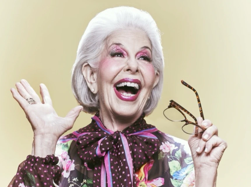 Elderly woman with white hair, makeup, floral top, holding glasses in photo studio smiling with hand raised in front of a yellow backdrop.